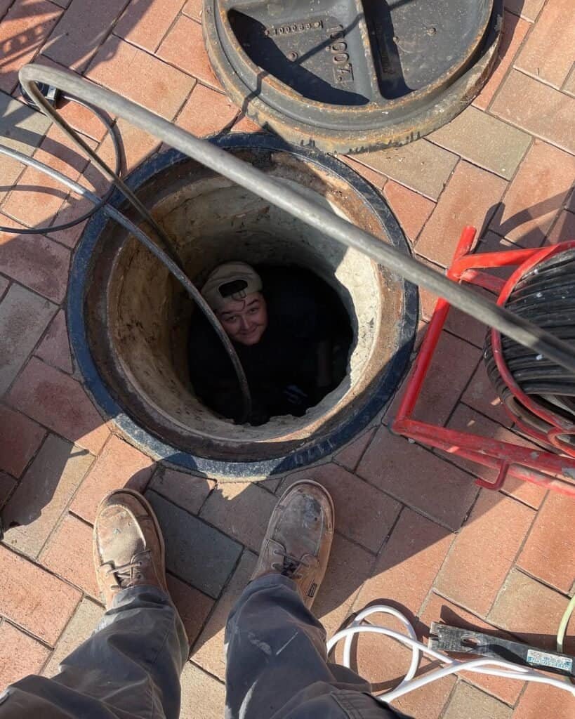 A Thompson Trenchless and Hydro Jetting worker inspects pipes in a manhole while another team member assists above.